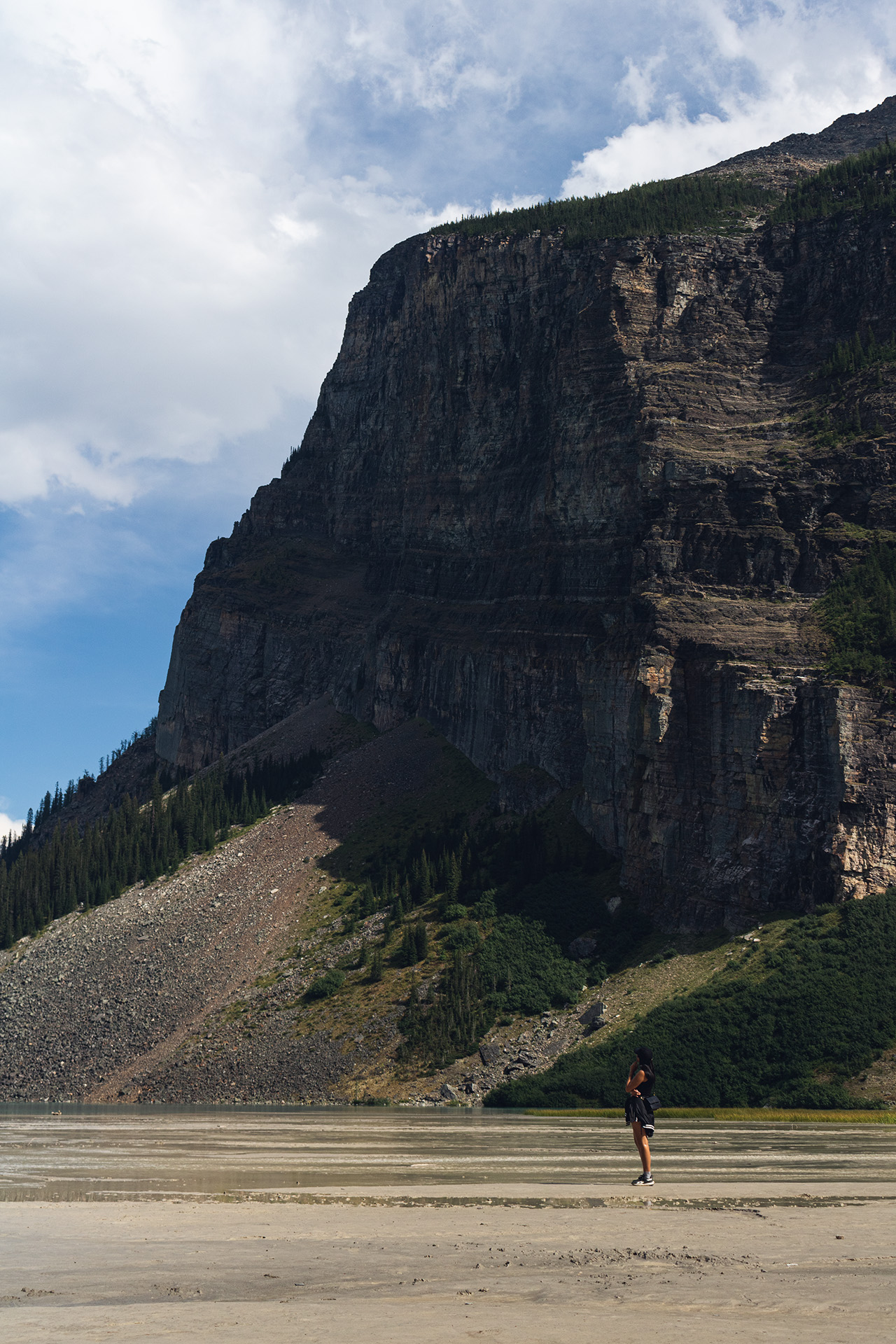 Girl standing on sandy dirt with shallow puddles of water and a giant rocky mountain behind her.
